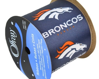 May include: A spool of navy blue ribbon featuring the Denver Broncos logo in white and orange. The ribbon is labeled "Offray" and includes text in multiple languages, including "Ribbon" and "Cinta."