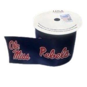 以下が含まれることがあります： 赤色の筆記体で「Ole Miss Rebels」と書かれた、濃い紺色のリボンのロール。リボンは部分的にほどかれ、テキストが表示されています。スプールは白で、印刷されたラベルが付いています。