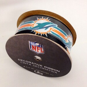 May include: A roll of decorative ribbon featuring the Miami Dolphins logo and the NFL logo. The ribbon is black with orange and teal accents. The text on the label reads "Decorative Ribbon Cinta Decorativa 2012 NFL Properties, LLC."