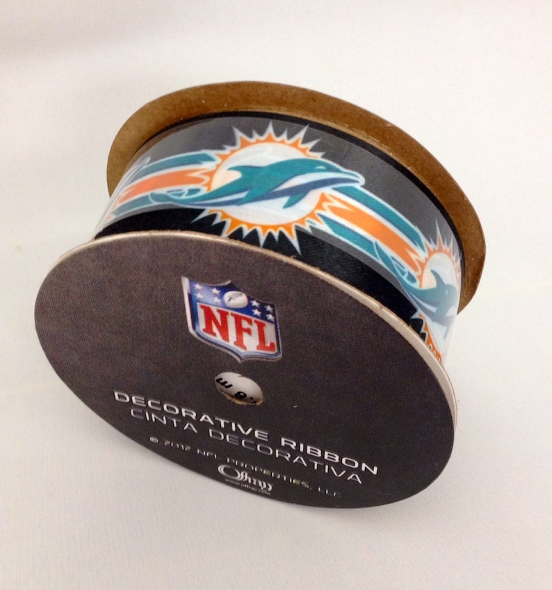 May include: A roll of decorative ribbon featuring the Miami Dolphins logo and the NFL logo. The ribbon is black with orange and teal accents. The text on the label reads "Decorative Ribbon Cinta Decorativa 2012 NFL Properties, LLC."