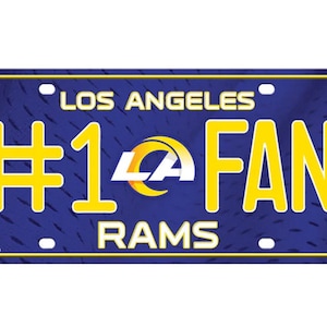 Puede incluir: Una placa de matrícula de los Los Angeles Rams azul y amarilla con el texto "#1 FAN" y el logotipo de los Rams. La placa tiene un fondo texturizado.
