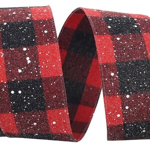May include: A wide fabric ribbon featuring a red and black buffalo plaid pattern. The surface of the ribbon is decorated with a scattered white speckled design that resembles falling snow, creating a textured appearance across the entire length.