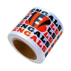 Könnte beinhalten: Eine Rolle weißes Band mit einem sich wiederholenden Muster des Wortes BENGALS in schwarzer und orangefarbener Schrift sowie einem großen orange-schwarzen B-Logo mit Tigerstreifen. Das Band hat eine gewebte Textur und ist auf einen Pappkern gewickelt.