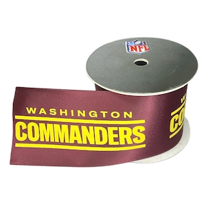 May include: A spool of burgundy ribbon featuring the words "WASHINGTON COMMANDERS" in yellow block letters. The ribbon has an NFL logo sticker on the top of the spool. The ribbon is unrolled, showing the team name.
