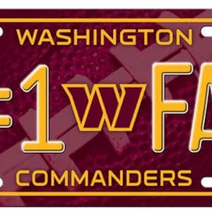 May include: A burgundy and gold license plate with the text "WASHINGTON #1 FAN COMMANDERS". The plate has a gold border and the team's logo in the center. The background has a subtle pattern.