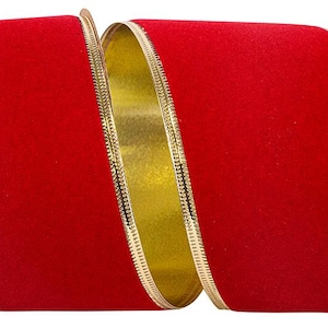 May include: A wide roll of red velvet ribbon featuring a smooth, deep red texture on the front surface and a metallic gold finish on the reverse side, with thin, textured gold metallic edges lining both sides of the ribbon throughout its length.