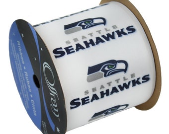 Puede incluir: Carrete de cinta blanca con el logotipo y el nombre de los Seattle Seahawks en azul marino y gris. La cinta presenta un patrón repetido del logotipo del equipo, una cabeza de halcón estilizada y el nombre del equipo. Fabricado por Offray.