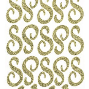 May include: A sheet of gold glitter stickers featuring fifteen individual letter S characters in a cursive, swirling script font, arranged in three rows of five, accompanied by four small gold glitter heart-shaped stickers at the bottom.