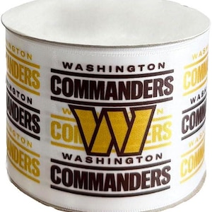 May include: A roll of white ribbon printed with the words "WASHINGTON COMMANDERS" in brown and gold. The ribbon features the team's logo, a gold "W" inside a brown outline. The ribbon is suitable for crafting and gift wrapping.