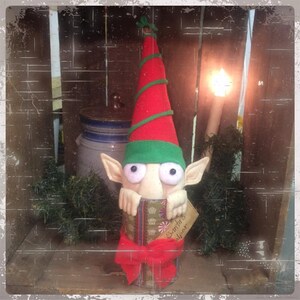 May include: A green and red elf doll peeking out of a wrapped gift box with a red bow. The gift box has a tag that says "Santa's Sleigh".