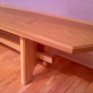 May include: A light wood bench with a simple design. The bench has a rectangular top with rounded edges and two legs on each side. The bench is made of natural wood and has a light brown finish.