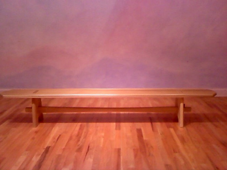 May include: A light wood bench with a simple design. The bench has a flat top and two legs on each end. The bench is sitting on a wooden floor.