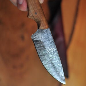 Pode incluir: Uma faca artesanal com cabo de madeira e l&acirc;mina de a&ccedil;o estampada. A l&acirc;mina tem um design &uacute;nico e rodopiante. O cabo &eacute; de cor castanho claro. A faca &eacute; segurada contra um fundo desfocado.