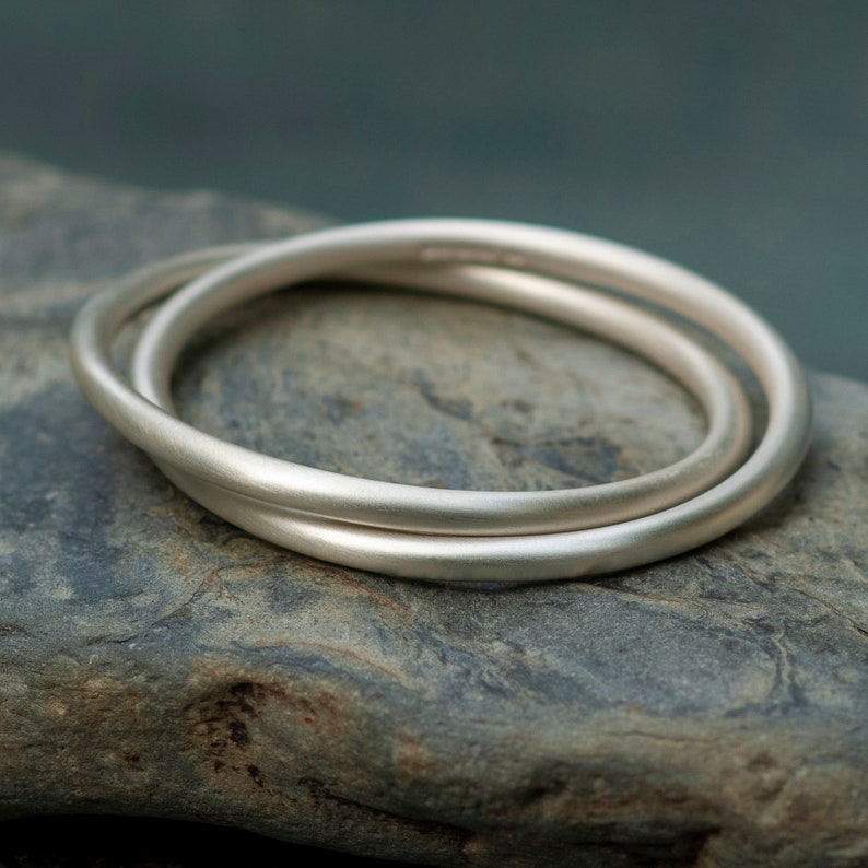 Op de afbeelding: Twee zilveren ringen, waarvan er &eacute;&eacute;n iets groter is dan de andere, zijn met elkaar verweven en liggen op een ruwe, grijze steen. De ringen hebben een gladde, geborstelde afwerking.