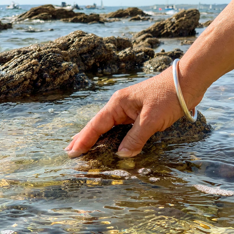 Op de afbeelding: Een hand reikt in het ondiepe water van een rotsachtig strand en raakt een gladde, natte rots. De hand draagt een zilveren armband.