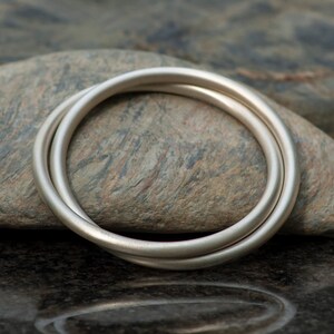 Op de afbeelding: Twee zilveren ringen, waarvan er &eacute;&eacute;n iets groter is dan de andere, zijn met elkaar verweven en liggen op een getextureerde grijze steen. De ringen zijn glad en gepolijst, met een subtiele glans.