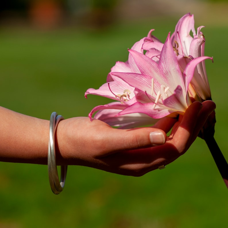 Op de afbeelding: Een hand die een roze bloem vasthoudt met witte accenten. De bloem heeft meerdere bloemblaadjes en heeft een delicate uitstraling. De hand draagt een zilveren armband.