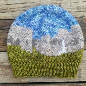 May include: A knitted hat with a landscape design. The hat is green at the bottom, with a brown and white mountain scene in the middle, and a blue sky at the top.