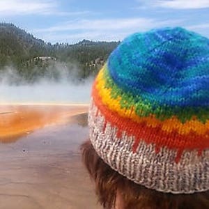 Puede incluir: Un gorro de punto colorido con un patrón de arcoíris y un borde blanco. El gorro lo lleva puesto una persona de pie frente a un paisaje natural.