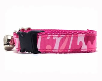 May include: A pink cat collar with a camouflage pattern in shades of pink and white. The collar has a black plastic buckle and a silver bell attached. The collar is designed for pets.
