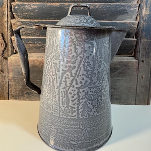 May include: A vintage gray enamelware coffee pot with a lid and handle. The pot has a textured surface and a spout for pouring. The handle is dark and the pot is set against a rustic wooden background.