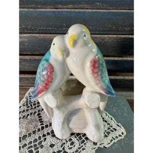 May include: A vintage ceramic bird figurine featuring two birds perched together on a branch. The birds are primarily white with accents of yellow, blue, and red. The figurine is a decorative item.