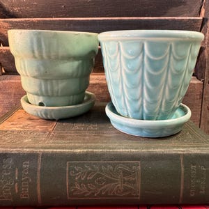 May include: Two ceramic plant pots with matching saucers. The left pot is a matte sage green with horizontal tiered ridges. The right pot is a glossy light turquoise blue featuring a vertical scalloped pattern around the exterior surface.