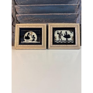 May include: Two framed silhouette art pieces. The frames are gold-colored with a decorative pattern. The art features black and white figures in a vintage style. One depicts a child blowing bubbles, the other a couple dancing.