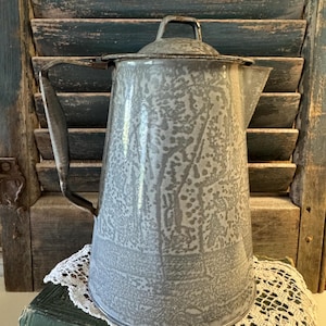 May include: A tall, tapered enamel coffee pot featuring a mottled gray and white speckled finish. The pot includes a matching lid with a small handle, a side handle, and a pouring spout. The surface has a smooth, glossy appearance with visible metallic edges along the rim and handle attachments.