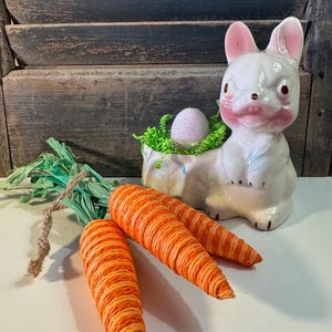 May include: A white ceramic bunny figurine with pink accents, holding a nest with a glittery egg. Three orange and yellow decorative carrots with green stems are in the foreground. The background features a weathered wooden shutter.