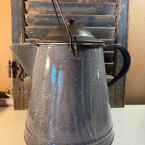 May include: A vintage-style, gray enamelware coffee pot with a black handle and lid. The pot has a spout and a wire handle for carrying. The background features a distressed blue shutter.