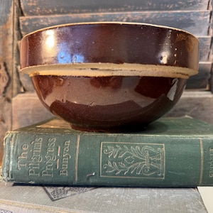 May include: A vintage brown stoneware bowl with a glossy finish. The bowl sits atop a green book titled "The Pilgrim's Progress" by Bunyan. The background features weathered, blue-gray wooden shutters, creating a rustic aesthetic.
