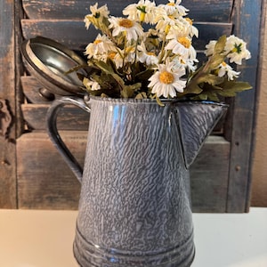 May include: A gray enamelware pitcher with a textured surface, filled with white and yellow daisy-like flowers. The pitcher has a handle and a spout, and the lid is partially open. The background features a weathered, blue wooden shutter.