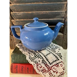 May include: A blue ceramic teapot with a lid and handle, set on top of a stack of books and a white lace doily. The teapot has a white rim and a curved spout. The background features a weathered, blue-painted wooden shutter.