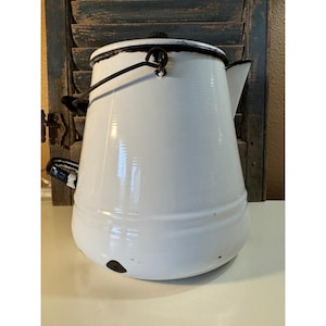 May include: White enamelware coffee pot with black trim and handle. The vintage pot has a rounded base and a lid with a black knob. The spout is on the right side. The pot is on a white surface with a blue shutter background.