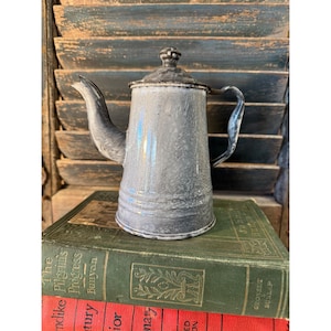 May include: A vintage, gray and white enamelware coffee pot with a lid and spout. The pot has a handle and sits on top of a stack of antique books with green and red covers. The background features weathered, blue wooden shutters.