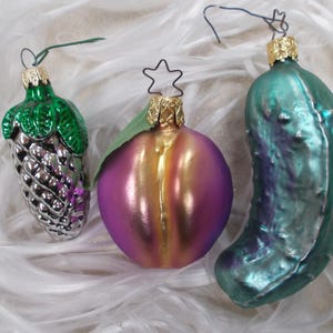 May include: Three glass Christmas ornaments: a silver pinecone with green leaves, a purple and gold fruit with a star topper, and a teal pickle. Each ornament has a gold cap and a string for hanging.