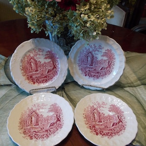 May include: A set of four white ceramic plates featuring a scalloped edge and a detailed crimson red transferware print depicting a landscape with architectural ruins, trees, and foliage. Each plate displays an identical scene in the center.