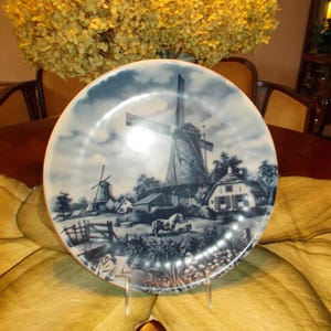 May include: A decorative plate featuring a windmill scene in shades of blue and white. The plate depicts a rural landscape with windmills, houses, and a cloudy sky. The plate is displayed on a clear stand.