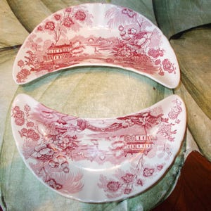 May include: Two crescent-shaped ceramic dishes featuring a detailed red and white transferware pattern. The design depicts a landscape with buildings, trees, and floral elements. The curved plates have a smooth, glossy finish and scalloped edges.