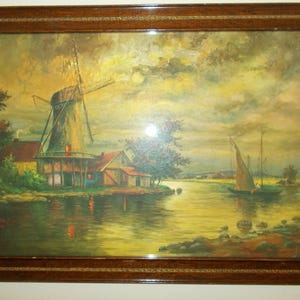 May include: A framed landscape painting depicting a windmill and buildings on a riverbank. The artwork features a windmill with turning blades, a sailboat, and a cloudy sky. The frame is a dark brown color.
