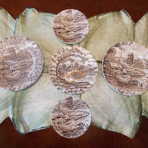 May include: A collection of brown and white ceramic plates with a pastoral scene design. The plates vary in size and are arranged on a green and white floral patterned surface. The plates feature images of houses, trees, and people.