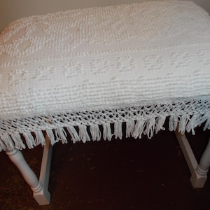 May include: A white rectangular bench featuring a textured, crocheted fabric cover with a geometric pattern. The cover includes a decorative crocheted fringe border with hanging tassels along the edges, supported by four white wooden legs.
