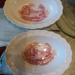 May include: Two oval ceramic serving bowls with a cream-colored base and a scalloped rim featuring embossed heart details. Each bowl displays a central maroon transferware illustration depicting a landscape with architectural ruins and trees.