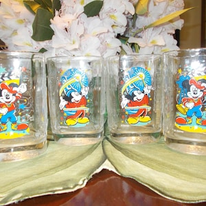 May include: A set of four clear glass tumblers featuring a colorful graphic of Mickey Mouse in a red and blue outfit with a yellow hat. Each glass displays the text 2000 and Disney, with a film clapperboard and star accents printed on the front.