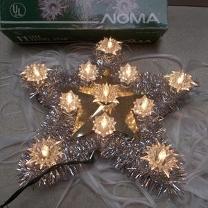 May include: A Noma 11-lite tinsel star tree topper featuring a gold-toned centre star surrounded by silver metallic tinsel and eleven clear, star-shaped light covers. The product is shown with its original green box labelled Noma 11 Lite Tinsel Star.
