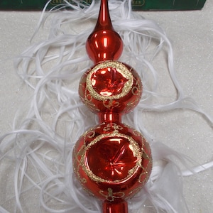 Può includere: Un puntale per albero di Natale in vetro rosso con un lungo elemento a punta e due sezioni sferiche sovrapposte. Ogni sfera presenta un design centrale a riflettore rientrante bordato di glitter oro e decorazioni.