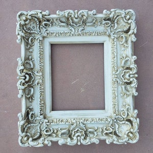 May include: A white ornate picture frame with a detailed, intricate design. The frame is made of wood and has a distressed finish.
