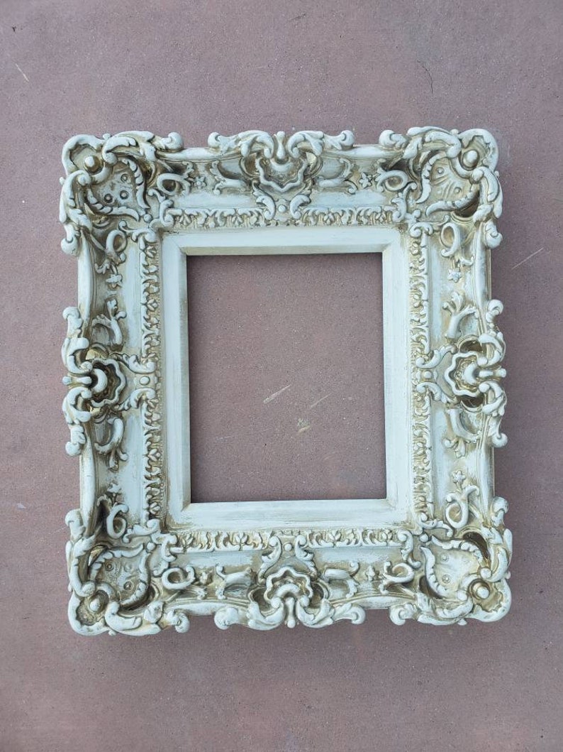 May include: A white ornate picture frame with a detailed, intricate design. The frame is made of wood and has a distressed finish.
