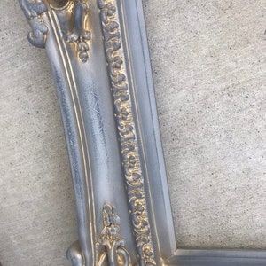 Puede incluir: Un marco de imagen adornado en plata y oro con un dise&ntilde;o detallado e intrincado. El marco est&aacute; hecho de madera y tiene un acabado desgastado.